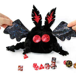 May include: A black plush bat-shaped dice bag with red eyes and wings. The bag is open and there are several dice spilling out of it, including red, brown, and gold dice.