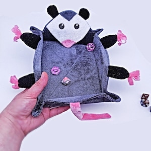 May include: A grey plush dice tray shaped like a possum with pink accents. The tray is open and has three dice inside.