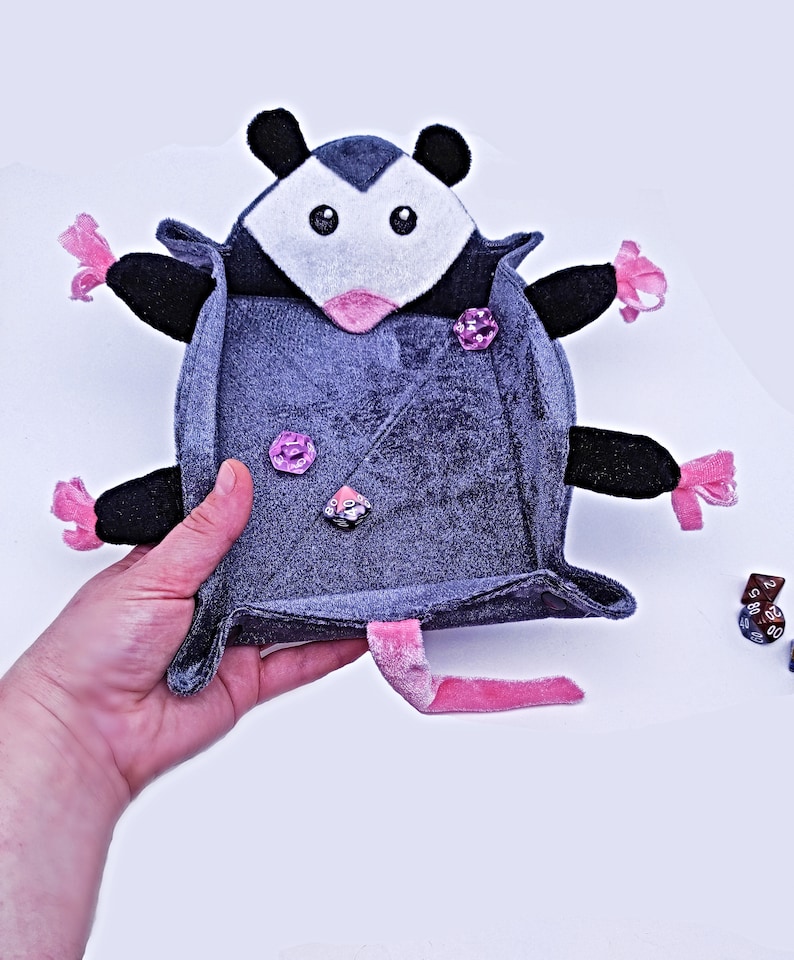 May include: A grey plush dice tray shaped like a possum with pink accents. The tray is open and has three dice inside.