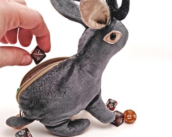 May include: A plush gray bunny with black horns and brown ears, holding a zippered pouch. The pouch is open and a hand is placing a black and white dice inside. There are several other dice on the floor, including a red and white dice, a brown and white dice, and a clear orange dice.