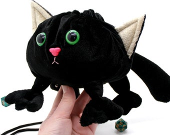 May include: Black plush cat-shaped dice bag with green eyes and a pink nose. The bag is open and has a drawstring closure. There are two green dice on the floor in front of the bag.