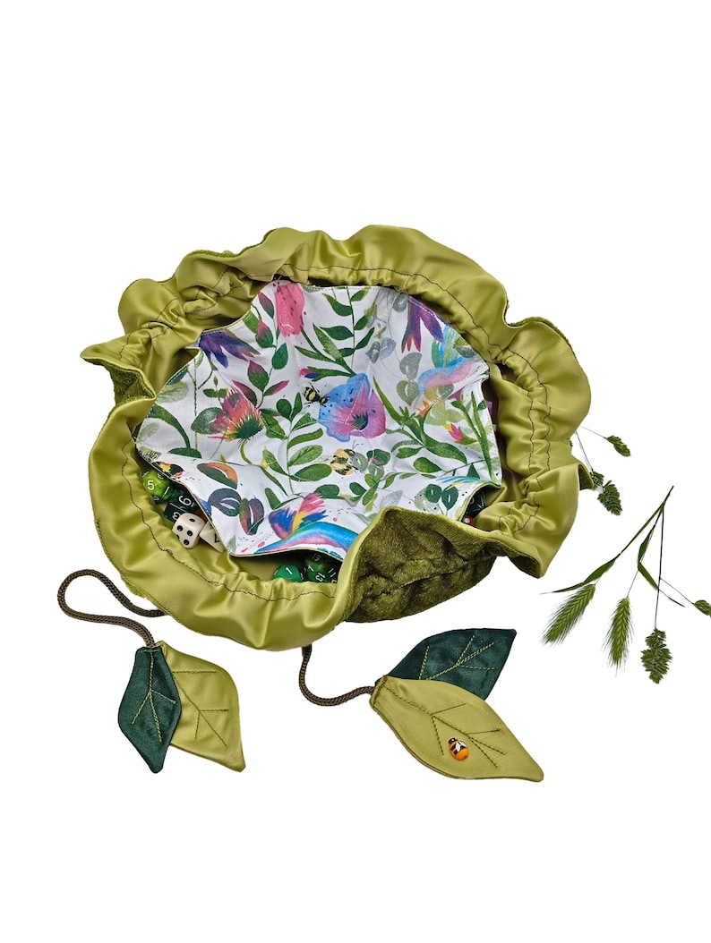 Puede incluir: Una bolsa de dados de terciopelo verde con cierre de cord&oacute;n y tiradores en forma de hoja. La bolsa est&aacute; forrada con una tela blanca con estampado floral con mariposas y abejas. La bolsa est&aacute; llena de dados verdes y blancos.
