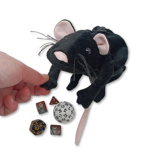 Pu&ograve; includere: Sacchetto da dado in peluche nero a forma di topo con naso e orecchie rosa. Il sacchetto &egrave; aperto e una mano ci sta entrando. Ci sono quattro dadi sul tavolo accanto al sacchetto, tra cui un d20, un d6, un d4 e un d100.