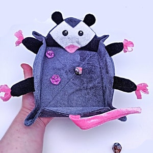 May include: A grey plush opossum-shaped dice tray with pink accents and a pink felt lining. The tray has three pink dice inside.