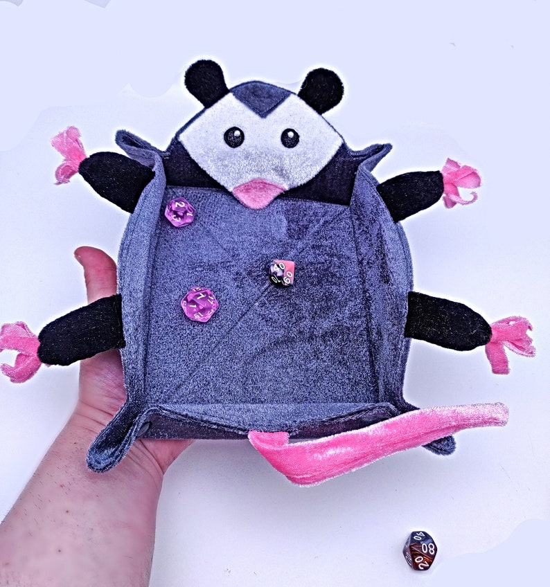 May include: A grey plush opossum-shaped dice tray with pink accents and a pink felt lining. The tray has three pink dice inside.