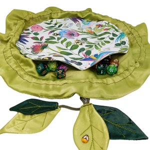 Puede incluir: Una bolsa de dados de tela verde con forma de flor con dos hojas verdes unidas. La bolsa est&aacute; abierta y llena de dados de colores.