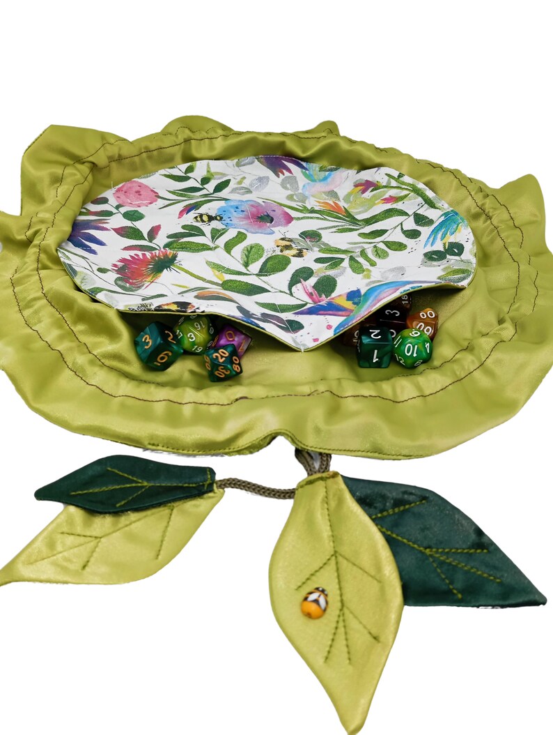 Puede incluir: Una bolsa de dados de tela verde con forma de flor con dos hojas verdes unidas. La bolsa est&aacute; abierta y llena de dados de colores.