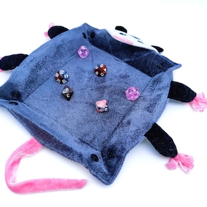 May include: A plush grey and black possum-shaped dice tray with a pink tail. The tray is filled with various coloured dice, including pink, red, and brown.