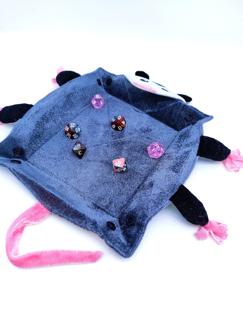 May include: A plush grey and black possum-shaped dice tray with a pink tail. The tray is filled with various coloured dice, including pink, red, and brown.