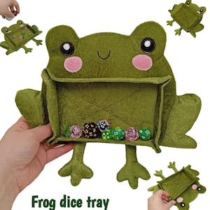 Pode incluir: Uma bandeja de dados em forma de sapo de feltro verde com bochechas rosa e olhos pretos. A bandeja contém vários dados de várias faces. O texto "Frog dice tray" está na parte inferior da imagem. Bandejas de sapo adicionais são mostradas.