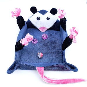 May include: A plush blue and black possum dice tray with pink felt accents. The tray has a pink felt tail and four dice are inside the tray.