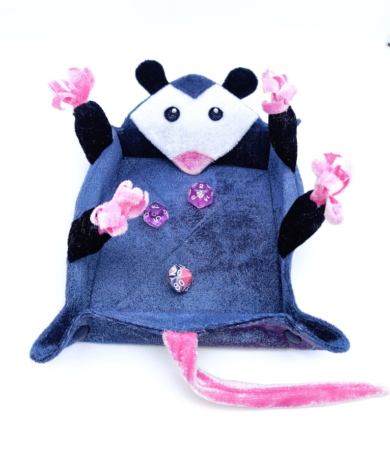 May include: A plush blue and black possum dice tray with pink felt accents. The tray has a pink felt tail and four dice are inside the tray.