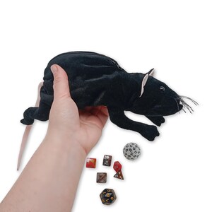 Pu&ograve; includere: Borsa da gioco in peluche nera a forma di topo con chiusura a zip. La borsa &egrave; tenuta in una mano e ci sono diversi dadi su una superficie bianca sottostante.