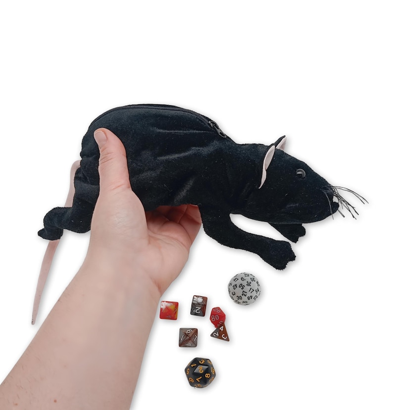 Pu&ograve; includere: Borsa da gioco in peluche nera a forma di topo con chiusura a zip. La borsa &egrave; tenuta in una mano e ci sono diversi dadi su una superficie bianca sottostante.