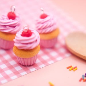 May include: Three pink and white cupcakes with a cherry on top. The cupcakes are on a pink and white checkered surface.