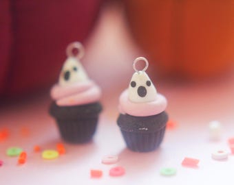 May include: Two miniature cupcake charms with ghost-shaped toppers. The cupcakes have black bases, pink icing, and white ghost faces with black dot eyes. Small colourful beads are scattered around the cupcakes.