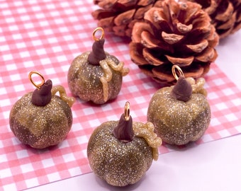 May include: Four miniature pumpkin charms with gold glitter, brown stems, and gold jump rings. The pumpkins are on a pink and white checkered surface, with brown pine cones behind them. Ideal for autumn-themed jewellery.