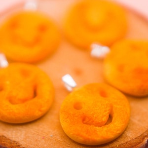 May include: Five orange smiley face charms on a wooden surface. The charms are made of a smooth, slightly textured material.