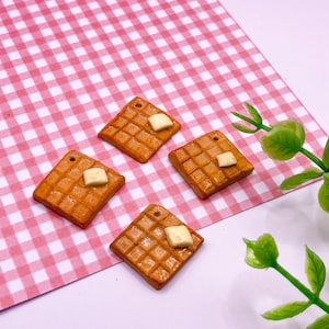 May include: Four miniature waffle charms with a square shape and a small pat of butter on top. The charms are on a pink and white gingham background.