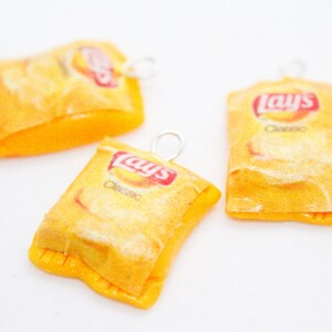 Puede incluir: Tres bolsas de patatas fritas Lay's en miniatura de color amarillo y rojo con anillas de salto plateadas. Las bolsas están hechas de arcilla polimérica y tienen el logotipo de Lay's y el texto "Classic" impreso.