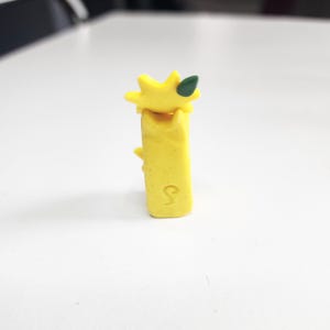 May include: A yellow figurine of a pineapple with a green leaf on top. The pineapple is standing upright on a white surface.