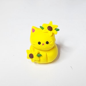 May include: A yellow cat figurine with a sunflower on its head and another sunflower in its paw. The cat has black eyes and a pink nose.