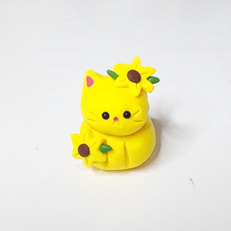 May include: A yellow cat figurine with a sunflower on its head and another sunflower in its paw. The cat has black eyes and a pink nose.