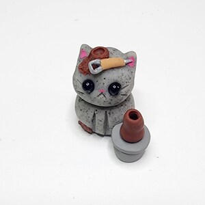 May include: A grey cat figurine with pink cheeks and black eyes, holding a small brown pot with a brown paintbrush on top. The cat is sitting on a white surface.