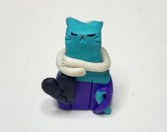 May include: A handmade cat figurine with a teal body, white arms, and purple trousers. The cat is hugging itself and has a broken black heart. The cat's tail is curled, and it has a red nose.