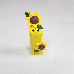 May include: A yellow clay figurine of a cat with two sunflowers on its head. The cat has black eyes and a brown nose.