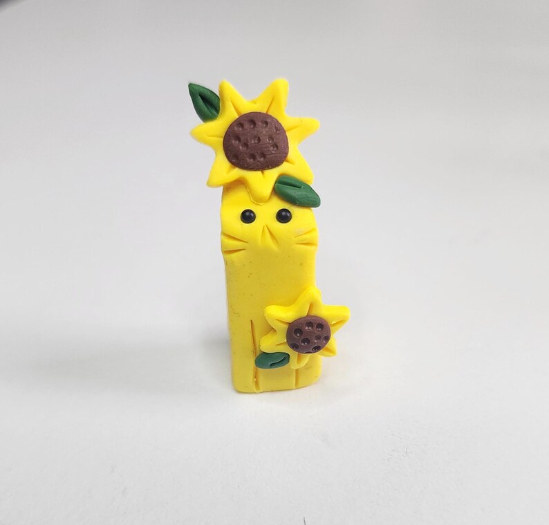 May include: A yellow clay figurine of a cat with two sunflowers on its head. The cat has black eyes and a brown nose.