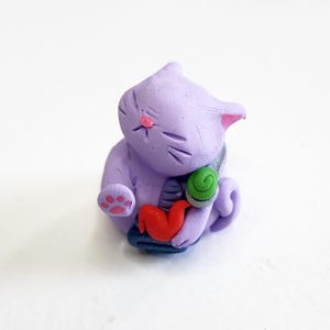 May include: A small purple cat figurine with a red, green, and blue swirl design. The cat is sitting with its paws tucked in and has a pink nose.