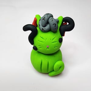 May include: A handmade green clay cat figurine with black question mark-shaped horns and a gray knot on its head. The cat has a pink nose, black eyes, and a curled tail. The figurine is set against a white background.