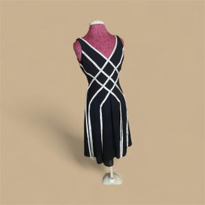 May include: A black sleeveless dress with white geometric accents. The dress features a V-neckline and a flared skirt, displayed on a pink dress form. The white accents create a lattice design across the bodice and down the skirt.