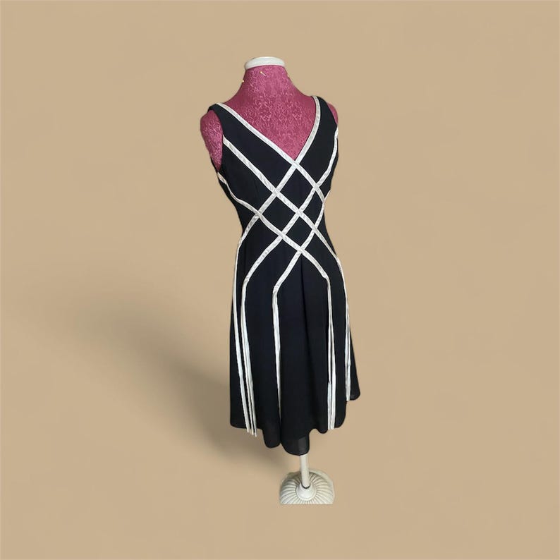 May include: A black sleeveless dress with white geometric accents. The dress features a V-neckline and a flared skirt, displayed on a pink dress form. The white accents create a lattice design across the bodice and down the skirt.