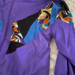 May include: A purple vintage windbreaker jacket with a full zipper and a hood. The jacket features color block panels in black, blue, and patterned fabric on the sleeves. The jacket is made of a shiny, lightweight material.