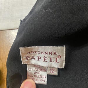 May include: Close-up of a black dress with a tag that reads "Adrianna Papell." The tag also indicates the dress is size 12 and made of 100% polyester.
