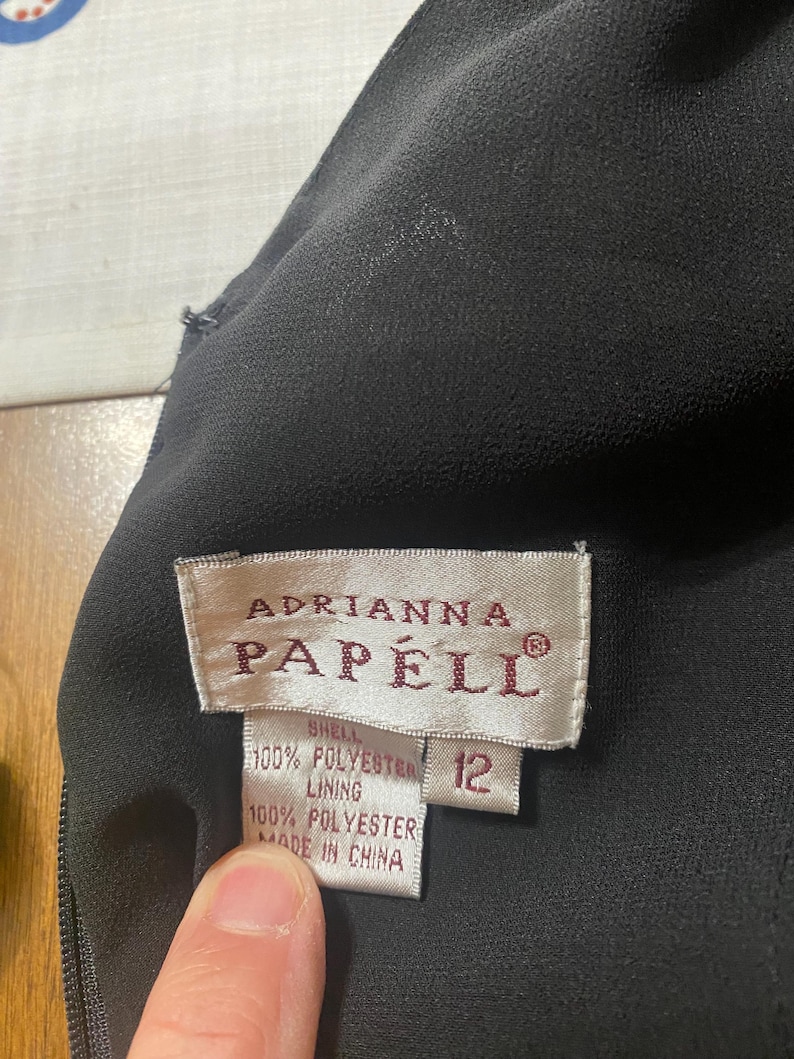 May include: Close-up of a black dress with a tag that reads "Adrianna Papell." The tag also indicates the dress is size 12 and made of 100% polyester.