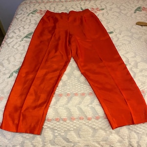 Puede incluir: Un par de pantalones de color naranja vibrante con un acabado suave y ligeramente brillante. Estos pantalones tienen una cintura estructurada, un diseño frontal limpio con sutiles pliegues verticales y piernas de corte recto hasta un dobladillo simple.