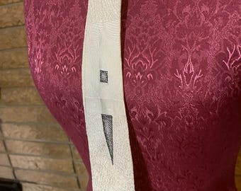 May include: Cream-colored fabric sash with geometric designs and a black triangle. The sash is displayed on a burgundy dress form with a floral pattern. The fabric has a textured appearance.
