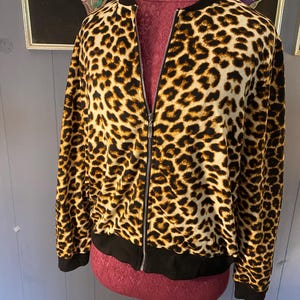 Puede incluir: Una chaqueta tipo bomber de manga larga con un estampado de leopardo en tonos marrón, negro y beige. La chaqueta tiene un cuello, puños y dobladillo acanalados en color negro. Una cremallera metálica plateada recorre verticalmente el centro.