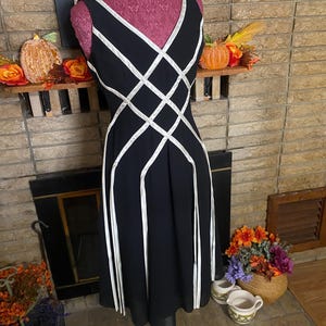 May include: Black sleeveless dress with white geometric accents. The dress features a V-neckline and a flared skirt. The dress is displayed on a pink dress form. The background includes a fireplace and decorative fall items.