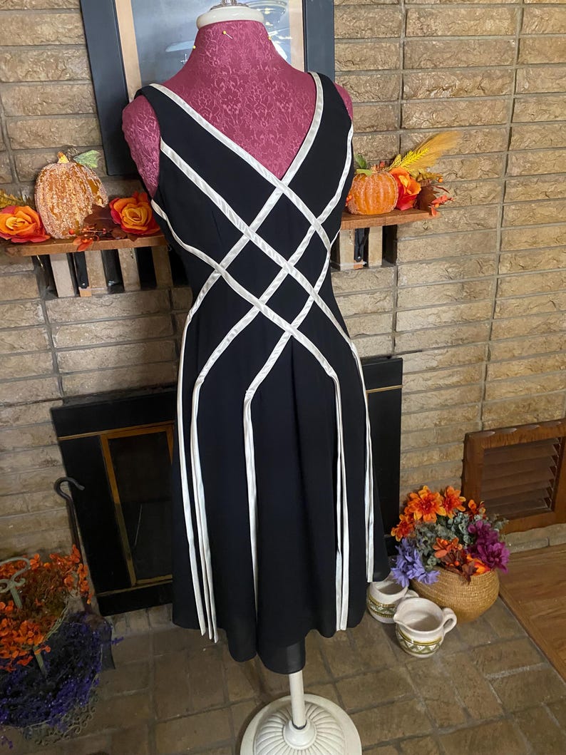May include: Black sleeveless dress with white geometric accents. The dress features a V-neckline and a flared skirt. The dress is displayed on a pink dress form. The background includes a fireplace and decorative fall items.
