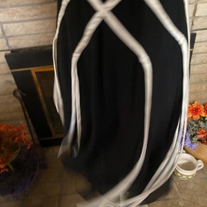 May include: A black dress with white crisscross ribbon detailing. The dress is displayed on a white mannequin base. The dress appears to be made of a flowing fabric.