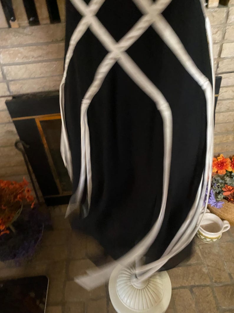 May include: A black dress with white crisscross ribbon detailing. The dress is displayed on a white mannequin base. The dress appears to be made of a flowing fabric.