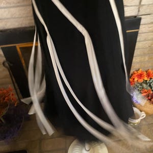 May include: A black dress with white ribbon accents. The dress is displayed on a white dress form. The ribbons flow down the dress, creating a flowing, elegant look. The dress is on a brick floor.