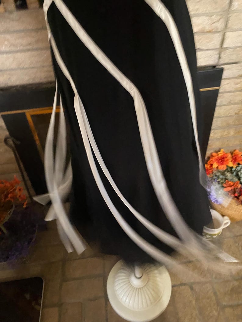 May include: A black dress with white ribbon accents. The dress is displayed on a white dress form. The ribbons flow down the dress, creating a flowing, elegant look. The dress is on a brick floor.
