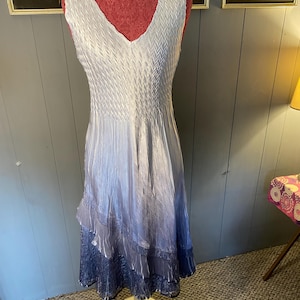 Puede incluir: Vestido sin mangas con cuello en V en un degradado de blanco a azul. El corpiño presenta un diseño plisado texturizado, que se transforma en una falda fluida con detalles en capas y con volantes. El vestido se exhibe en un maniquí.