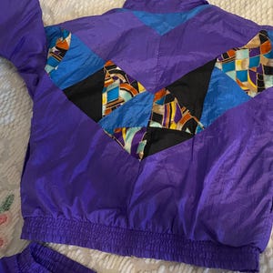 Könnte beinhalten: Eine violette Windjacke mit Stehkragen, langen Ärmeln und elastischem Saum. Das Rückenteil zeigt ein geometrisches Muster aus schwarzen, blauen und bunten abstrakten Formen, die in einem großen, spitz zulaufenden Chevron-Design angeordnet sind.