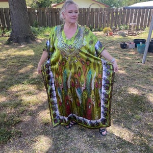 May include: A long, flowing green caftan with a V-neck and a peacock feather design. The caftan has a decorative neckline and a border of white and purple. The person is wearing black sandals.